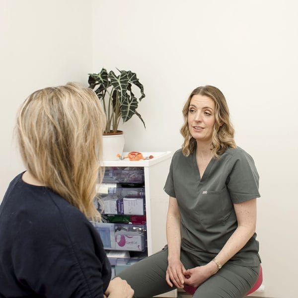 Two women engage in a Private Women's Health Consultation in a medical office; one in scrubs speaks while the other listens. A plant and medical supplies are visible in the background.